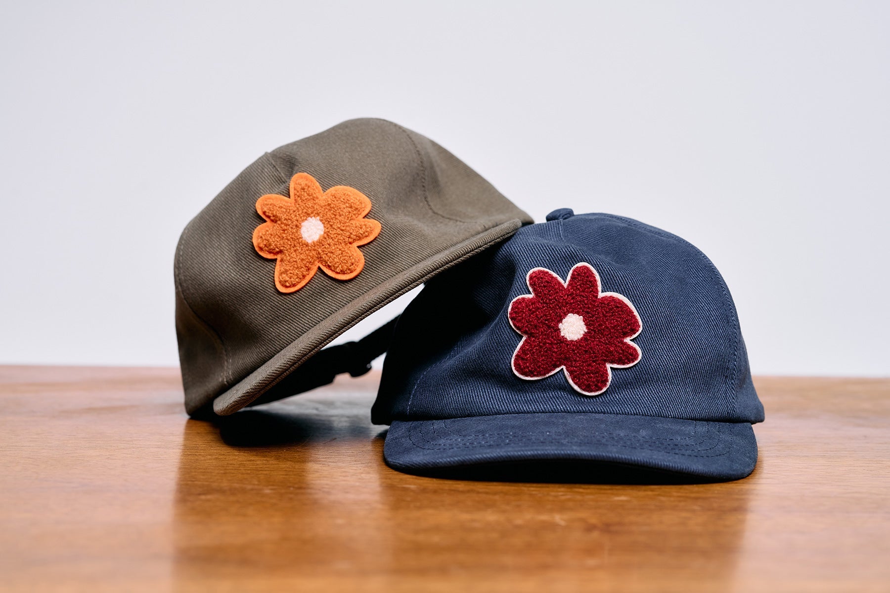 Two Wirth Hats featuring flower patches – one in olive with an orange flower and one in navy with a red flower, symbolizing self-care and the story behind the Flowers on Fridays hat.
